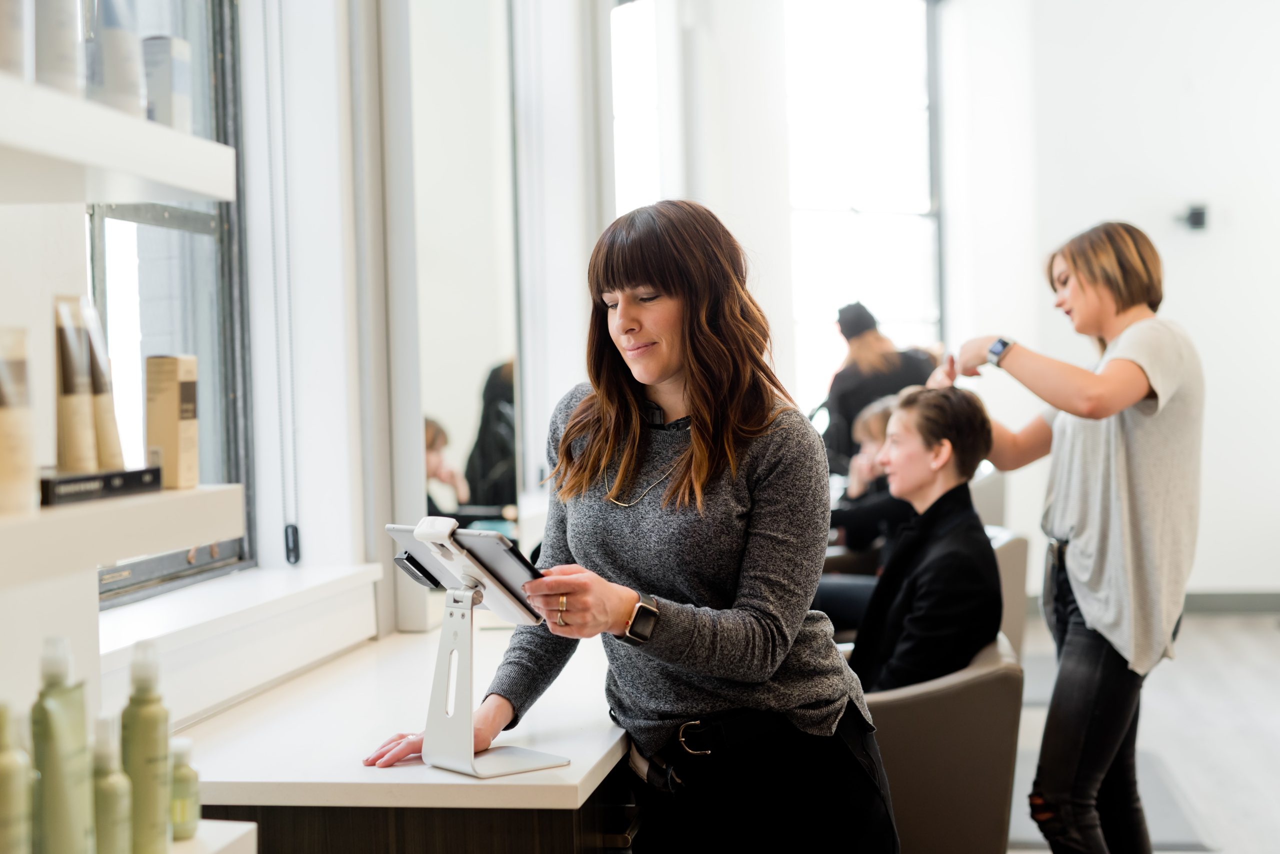 A woman with long brown hair uses a tablet at the salon counter, perhaps managing social media for this small business. Nearby, another woman with short hair styles a seated customers hair, and shelves filled with products line the bright, modern space.