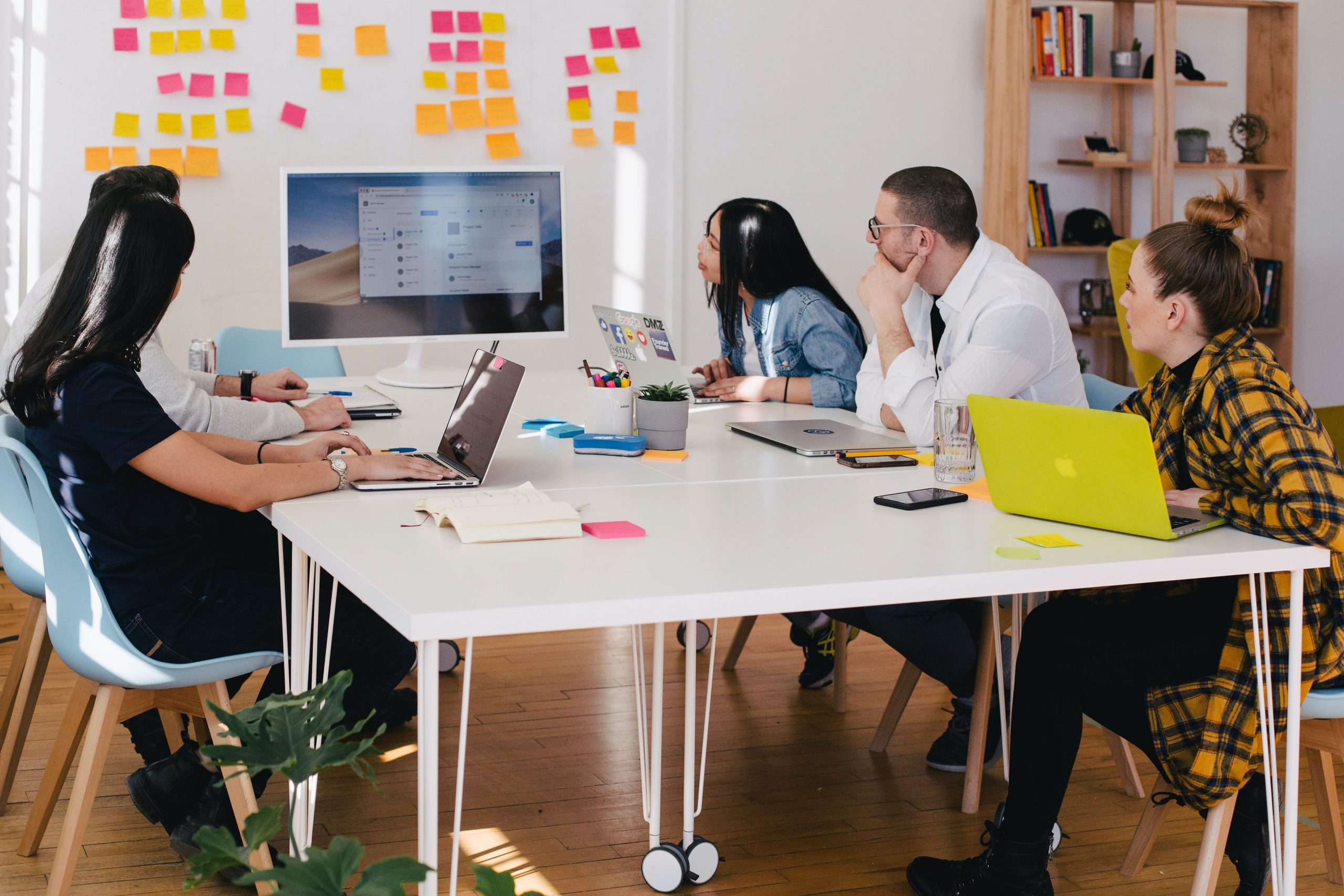 A group of five people sits around a white table, engaged in a lively discussion. Theyre analyzing a project on the computer screen, possibly conducting a SWOT analysis. Colorful sticky notes adorn the wall, while laptops and stationery clutter the table, each contributing to a stellar brainstorming session.