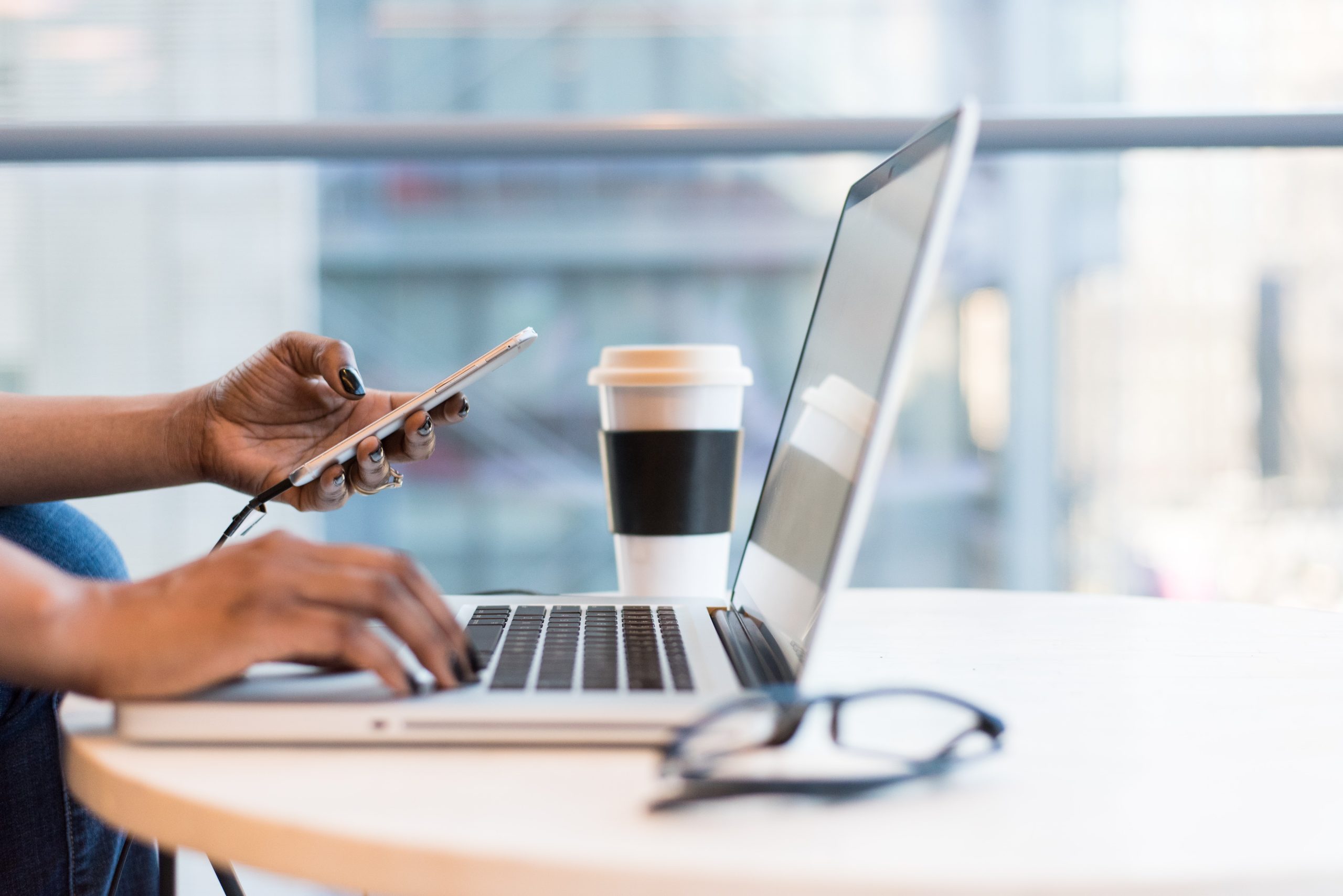 A person sits at a round table using a laptop, checking social media on a smartphone plugged into it. A coffee cup and glasses are also on the table. The background reveals large windows with an urban view, reflecting the bustling changes businesses face today.