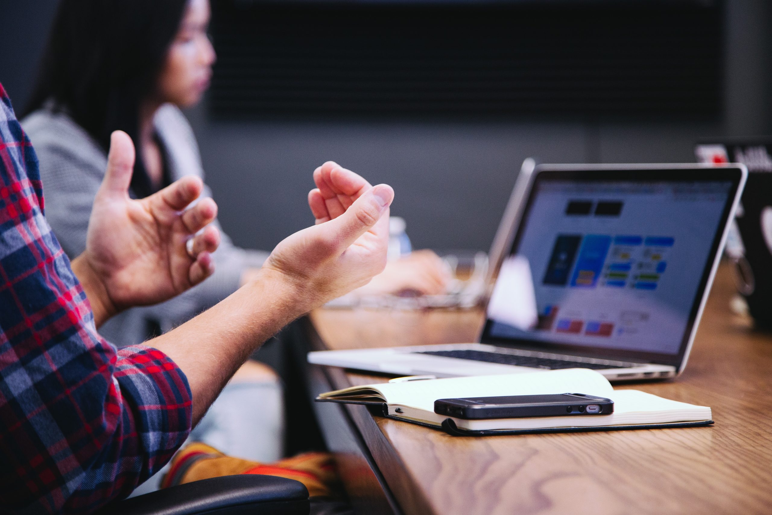 A person expertly gesturing with hands in front of a laptop, handling a social media crisis during a meeting. The screen displays graphs and data. Another individual is blurred in the background, while a notebook and smartphone rest on the wooden table.