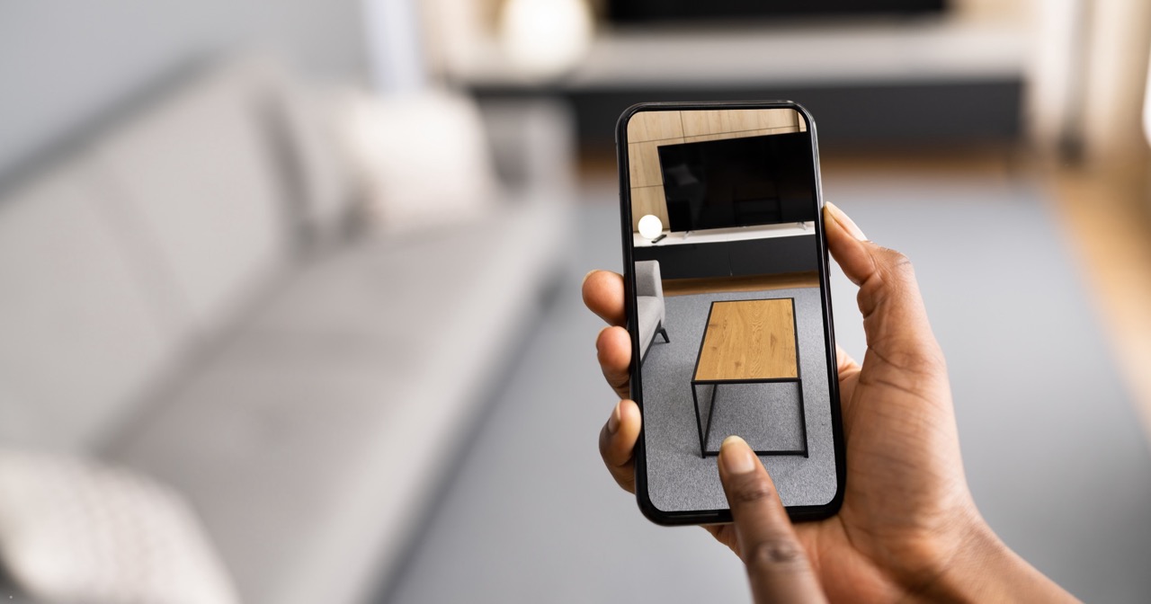 Close-up of a person holding a smartphone, using augmented reality to position a virtual wooden coffee table over a living room scene—an innovative touchpoint in digital marketing. A gray sofa and TV stand are visible in the blurred background.