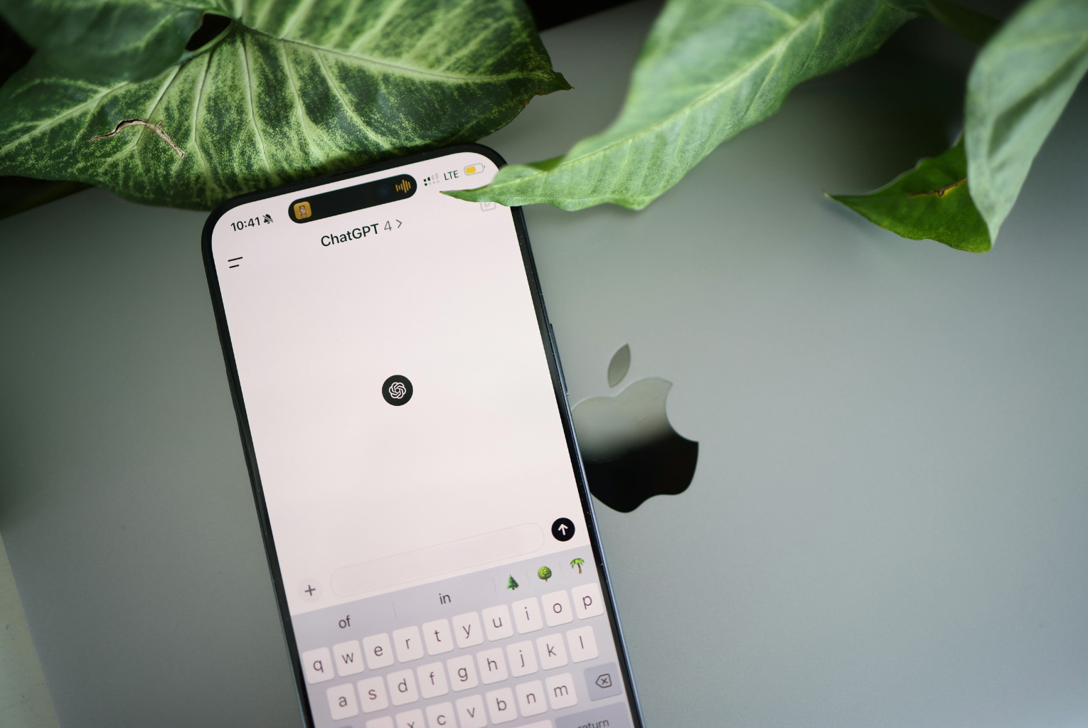 A smartphone displaying a chat interface with ChatGPT 4 AI is placed on a silver laptop with an Apple logo. The phone screen shows a text input area and a keyboard. Green leaves frame the devices, adding an organic touch to the tech scene.