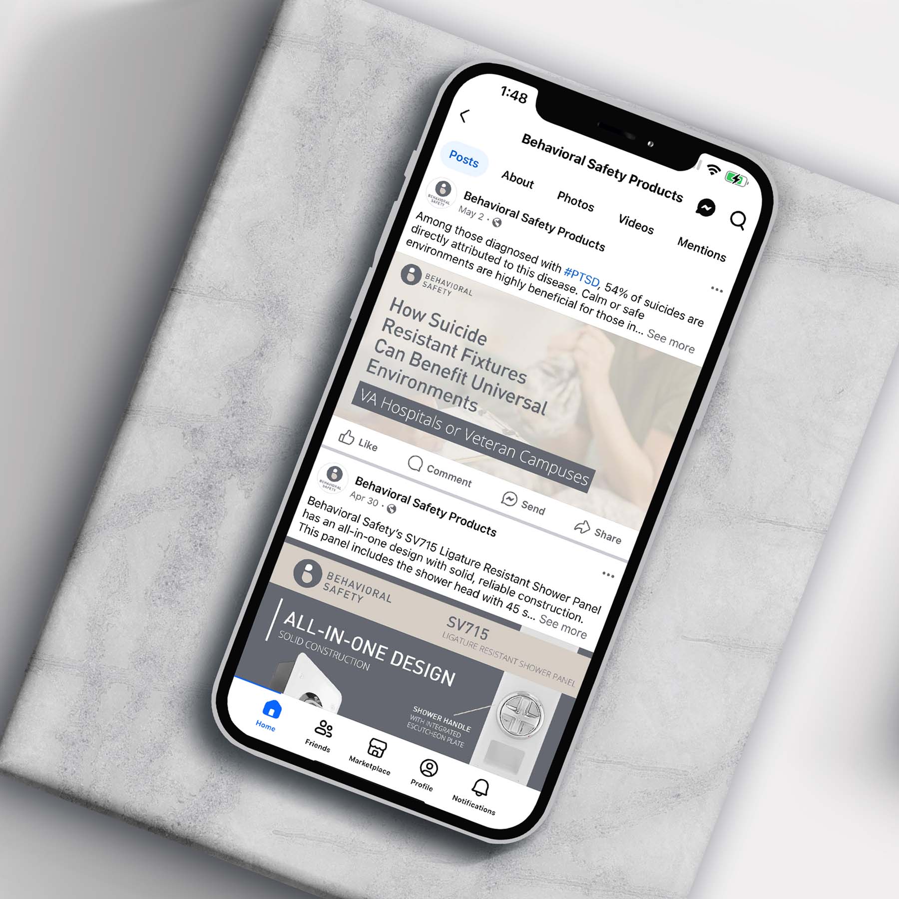 A smartphone displaying a Facebook page for Behavioral Safety Products. The page features a post about suicide-resistant fixtures in universal environments. The phone is on a textured gray surface.